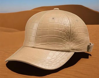 May include: A beige baseball cap with a crocodile skin effect. The cap features a curved peak and an adjustable strap. The background shows a desert landscape with sand dunes and a blue sky.