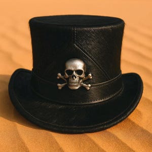 Puede incluir: Sombrero de copa negro con un emblema de calavera y huesos cruzados plateado. El sombrero tiene una copa redondeada y un ala ancha. El sombrero está hecho de un material texturizado y está sobre un fondo arenoso.