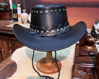 May include: A black leather cowboy hat with silver-colored studs along the brim and a decorative band. The hat has a wide brim and a chin strap. The hat is displayed on a wooden stand.