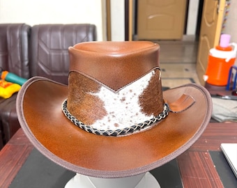 Op de afbeelding: Een bruine leren cowboyhoed met een wit en bruin patroon op de kroon. De hoed heeft een brede rand en een gevlochten zwarte band rond de basis van de kroon. Het wordt gepresenteerd op een witte standaard.