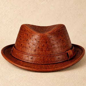 May include: A brown fedora hat with a textured surface. The hat has a structured crown, a wide brim, and a band around the base. The hat is made of a leather-like material. The hat's dimensions are not visible.