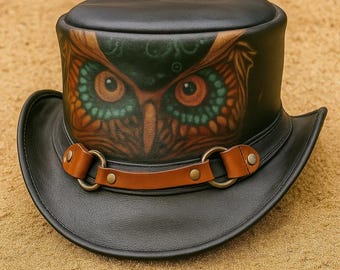 Puede incluir: Sombrero de copa de cuero negro con una banda de cuero marrón y anillos color latón. El sombrero presenta una ilustración detallada de un búho en tonos marrones, naranjas y verdes. El sombrero tiene un ala ancha y una copa redondeada.