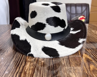 Puede incluir: Sombrero de vaquero con estampado de piel de vaca. El sombrero presenta un fondo blanco con manchas negras, una banda de cuero negro y un detalle de botón plateado. Un sombrero de estilo western clásico.