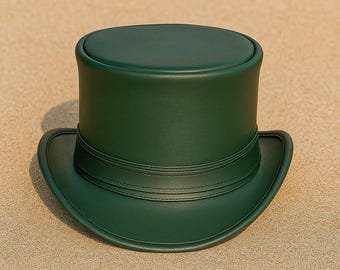 Puede incluir: Un sombrero de copa verde oscuro con una corona redondeada y un ala ancha. El sombrero presenta una banda alrededor de la base de la corona. El sombrero está hecho de un material liso y descansa sobre una superficie arenosa.