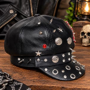 Puede incluir: Una gorra de cuero negro adornada con púas de metal plateado, tachuelas en forma de estrella, monedas circulares y pedrería transparente. La visera presenta ojales metálicos y una correa de cuero con tachuelas y una moneda central en la base.