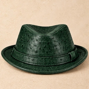 Op de afbeelding: Een donker bosgroene fedora hoed met een getextureerd oppervlak met kleine, verhoogde cirkelvormige patronen die doen denken aan struisvogelleer. De hoed heeft een bijpassende groene band rond de basis van de kroon en een gestructureerde rand.