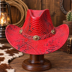 Puede incluir: Un sombrero de vaquero de color rojo brillante con un patrón de piel de serpiente texturizado y detalles en negro. El sombrero incluye una banda roja a juego adornada con tres tachuelas circulares de metal dorado colocadas simétricamente.