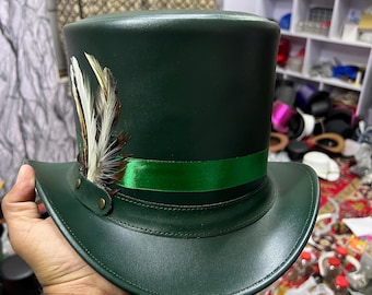 Puede incluir: Un sombrero de copa verde oscuro con una cinta verde y adornos de plumas. El sombrero tiene un ala ancha y una copa alta y cilíndrica. Las plumas son blancas y marrones. El sombrero lo sostiene una persona.