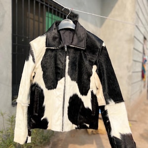 May include: A black and white cowhide jacket with a zip fastening. The jacket has a black collar and is suspended on a hanger. The design of the jacket incorporates patches of black and white fur.