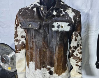 May include: A jacket featuring a brown and white cowhide pattern with a textured hair-on-hide finish. The garment includes a pointed collar, a front button closure with metallic buttons, and two chest pockets with buttoned flaps on the front.