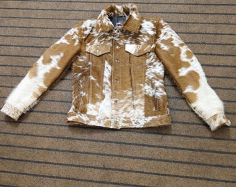 May include: A long-sleeve jacket featuring a textured cowhide pattern in shades of brown and white. The garment includes a pointed collar, a front button-down closure, two chest pockets with buttoned flaps, and long sleeves with buttoned cuffs.