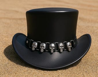 May include: A black hat with a wide brim and a tall, cylindrical crown. A silver band with skull accents encircles the hat. The hat is resting on a sandy surface, suggesting an outdoor setting. The hat combines elements of a top hat and a cowboy hat.