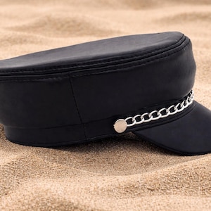 May include: Black hat with a short brim and a silver chain detail. The hat has a flat top and a structured shape. The hat is sitting on a sandy surface.