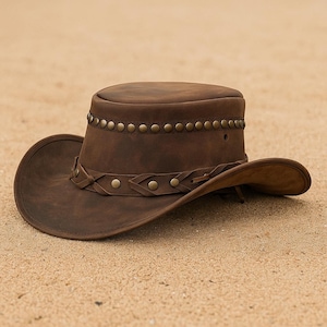 May include: A brown leather cowboy hat with a wide brim and a rounded crown. The hat features a decorative band with metal studs around the crown and a braided leather band. The hat is resting on a sandy surface.