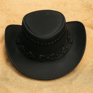 May include: A black leather cowboy hat with a wide brim and a rounded crown. The hat has a decorative band with rivets and braided leather accents around the crown. The hat is on a sandy surface.