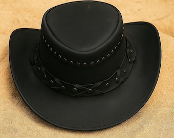 May include: A black leather cowboy hat with a wide brim and a rounded crown. The hat features a decorative band with rivets and braided leather accents around the crown. The hat is sitting on a sandy surface.