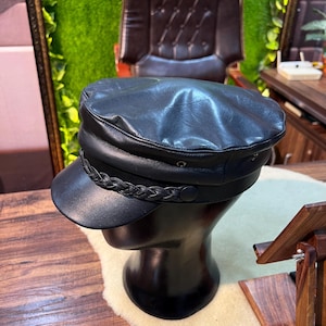 May include: A black leather cap with a braided band detail and a short brim. The cap is displayed on a black mannequin head. The hat has a classic, nautical style.