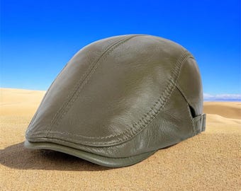 May include: Olive green leather flat cap with a curved brim. The cap has a stitched seam detail across the top and a small adjustable strap at the back. The hat is set against a desert backdrop with a blue sky.
