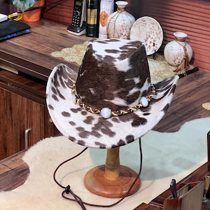 Op de afbeelding: Cowboyhoed met een bruin en wit gevlekt patroon, tentoongesteld op een houten standaard. De hoed heeft een decoratieve band met witte stenen en een bruine kinriem. Een accessoire met een western-thema.