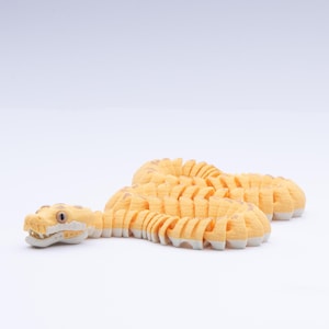 May include: A yellow and tan articulated snake toy. The toy snake has a detailed head with open jaws and a segmented body. The snake is lying on a white surface.