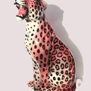 May include: A ceramic leopard figurine in a seated position with its mouth open, revealing white teeth and a pink interior. The body is finished in a glossy pink glaze with black leopard spots covering the head, neck, back, and legs.
