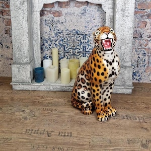 May include: A ceramic leopard figurine with a yellow and orange coat featuring black spots. The leopard is depicted in a seated position with its mouth open, revealing pink gums and white teeth. The eyes are green with black pupils.