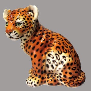 May include: A ceramic figurine of a seated leopard cub with a glossy orange coat covered in black spots. The cub has green eyes, white accents on the ears and muzzle, and a detailed facial expression. The sculpture is rendered in a realistic style.