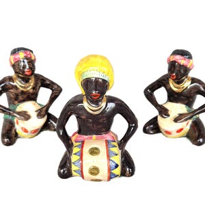 Peut inclure: Trois figurines en céramique représentant des personnes à la peau foncée, agenouillées et jouant du tambour. La figure centrale porte une coiffe jaune, tandis que les deux autres portent des bandeaux roses et jaunes. Toutes portent des colliers dorés.