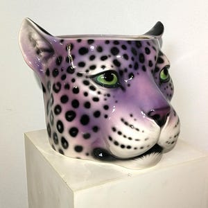 May include: A ceramic planter shaped like a leopard head, featuring a vibrant purple base color with black spots. The feline face has detailed green eyes, a dark nose, and white accents around the muzzle and ears, with an open top for plants.