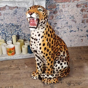 May include: A ceramic leopard statue, seated with its mouth open, showing teeth. The statue has a tan and black spotted pattern, green eyes, and a white underbelly. The background includes candles and a distressed brick wall.