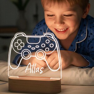 May include: A white illuminated video game controller night light with the name "Atlas" on it, set on a wooden base. The boy in the image is wearing blue pajamas. The light is on a bed.