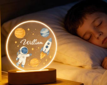 May include: A round, illuminated night light with a space theme, featuring an astronaut, rocket, and planets. The name "William" is written in cursive. The light sits on a wooden base, next to a sleeping child in a bed.