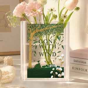 May include: A clear rectangular acrylic vase featuring a printed graphic of a tree, green landscape, and the text GONE WITH THE WIND by MARGARET MITCHELL. The vase contains a bouquet of light pink flowers, white blossoms, and green stems.