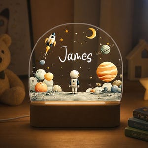 May include: A personalized night light featuring a space-themed design with a rocket, planets, and an astronaut. The name "James" is written in script. The light sits on a wooden base and is surrounded by a teddy bear and books.