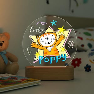 May include: A personalized night light featuring a cartoon cat design with the name "Poppy" in blue and "Evelyn" in white. The light has a wooden base and a clear acrylic panel with a star-shaped outline. The cat is orange and white.