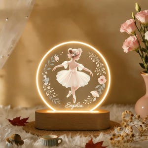 Könnte beinhalten: Ein rundes, beleuchtetes Nachtlicht mit einer Ballerina-Illustration. Das Design zeigt eine Ballerina in einem Tutu, umgeben von floralen Akzenten, mit dem Namen "Sophia" darunter. Das Licht steht auf einem Holzsockel.