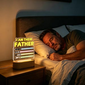 May include: A night light with the text "I AM THEIR FATHER" and illustrations of lightsabers. The light has a wooden base and a clear acrylic panel with the text and lightsaber designs. The lightsabers glow in various colors. The light is on a nightstand next to a bed.