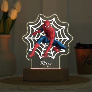 May include: A Spider-Man themed night light. The light features a red and blue Spider-Man figure in front of a white spiderweb design. The name "Riley" is written on the base. The light sits on a wooden base.