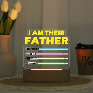 May include: A lighted acrylic sign with the text "I AM THEIR FATHER" in yellow. Below are three lightsaber designs with blue, pink, and green beams, each with a name. The base is a wooden block.