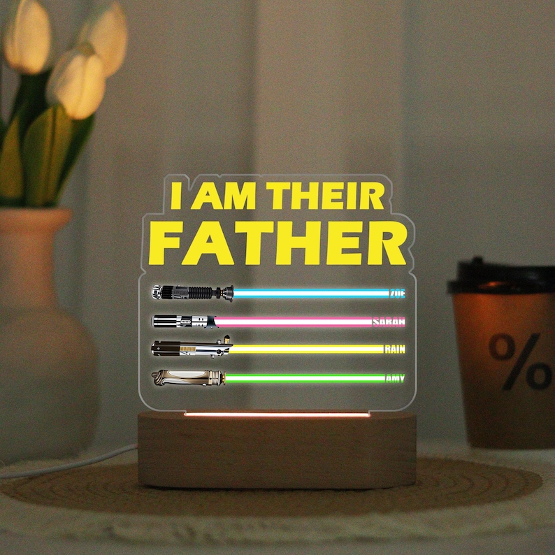 May include: A lighted acrylic sign with the text "I AM THEIR FATHER" in yellow. Below are three lightsaber designs with blue, pink, and green beams, each with a name. The base is a wooden block.