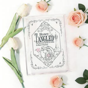 Könnte beinhalten: Transparente Kunststoffhülle mit dem Disney-Film "Tangled". Das Cover zeigt den Titel, eine Illustration von Rapunzel und den Text "Der Weg zu deinem Schicksal liegt in der Magie deines Herzens". Die Hülle ist von rosa Rosen und weißen Tulpen umgeben.