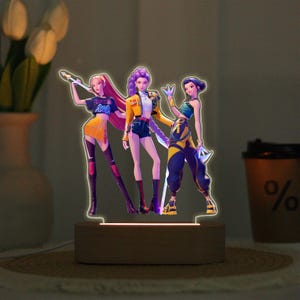 May include: An LED lamp with three stylized figures in colourful outfits. The figures have a glowing outline and are mounted on a wooden base. The lamp emits a warm light, creating a modern and artistic aesthetic, suitable for a bedroom.