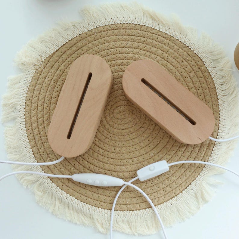 May include: Two light-colored wooden LED lamp bases with a long, narrow slot for holding a light. The bases are connected by white cords with a switch. The lamps sit on a round, woven mat with a fringed edge.