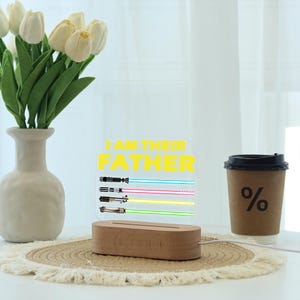 May include: A decorative light with the text "I AM THEIR FATHER" in yellow. The light features four lightsaber designs with blue, pink, yellow, and green beams. The base is made of wood. A coffee cup and vase of flowers are also in the image.