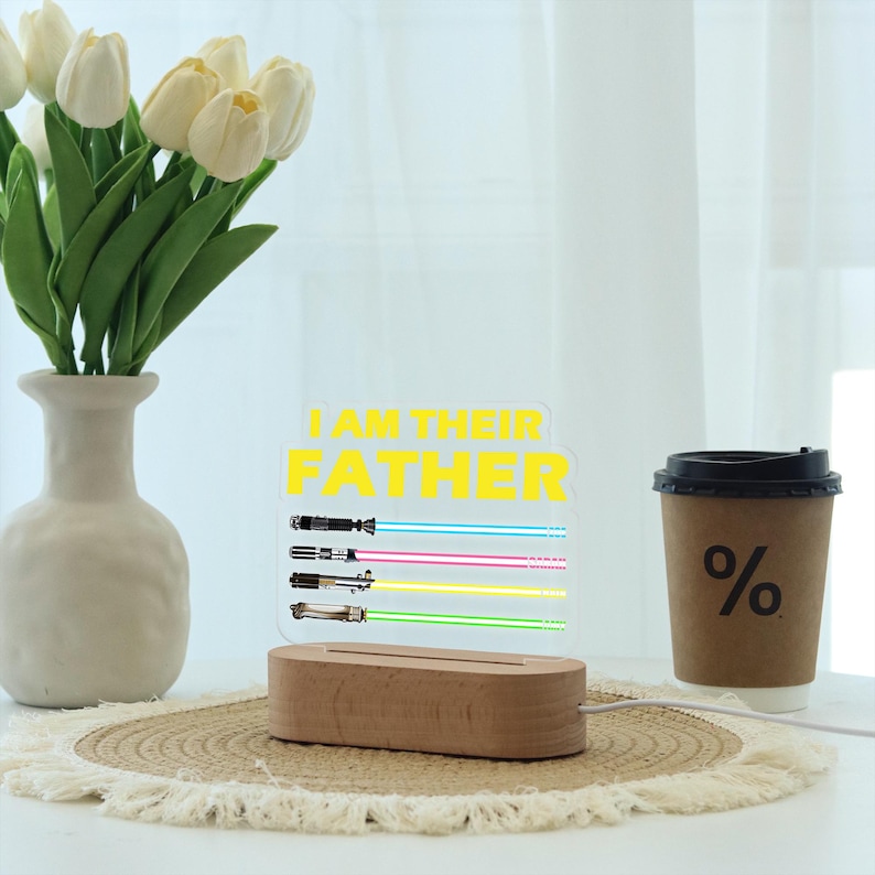 May include: A decorative light with the text "I AM THEIR FATHER" in yellow. The light features four lightsaber designs with blue, pink, yellow, and green beams. The base is made of wood. A coffee cup and vase of flowers are also in the image.