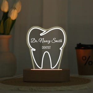 Può includere: Lampada a LED luminosa a forma di dente, con il nome "Dr. Nancy Smith" e la parola "DENTIST" incisi. La lampada è posizionata su una base rettangolare in legno. Lo sfondo include un vaso con fiori bianchi e una tazza di caffè.