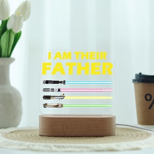 May include: A clear acrylic night light with the text "I AM THEIR FATHER" in yellow. Below the text are three lightsaber designs with blue, pink, yellow, and green beams. The light sits on a wooden base.
