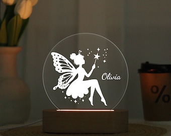 May include: A decorative night light featuring a fairy design. The fairy is white and has butterfly wings, holding a wand with stars. The name "Olivia" is written on the clear, circular light. The base is made of wood.