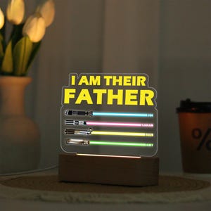 May include: A lighted acrylic sign with the text "I AM THEIR FATHER" in yellow. Below are three lightsaber designs with names: Zoe, Sarah, Rain, and Amy. The sign is set on a wooden base.