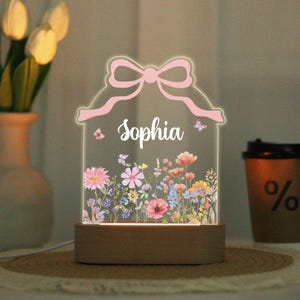 Op de afbeelding: Gepersonaliseerd nachtlampje met de naam "Sophia" in wit verlicht. Het acryl paneel heeft een roze strik ontwerp en een kleurrijke bloementuin. De basis is van hout. Vlinders zijn ook in het ontwerp opgenomen.